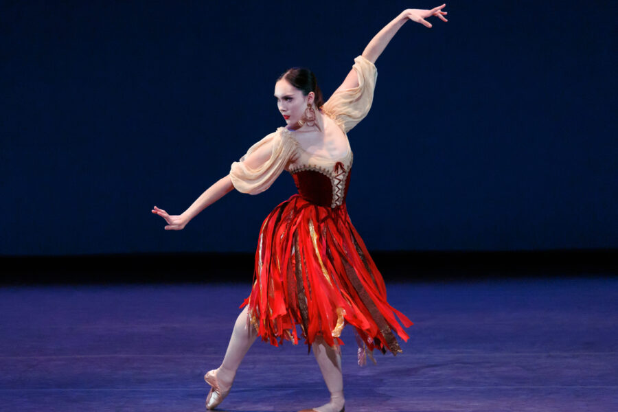 Mira Nadon on pointe in a red ribbon skirt, one arm lifted overhead, against a deep blue backdrop.