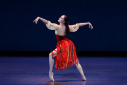 Mira Nadon on pointe in a wide lunge, arms extended and one knee raised, red ribbon skirt swinging.