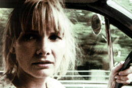 A blonde woman drives a car, one hand on the wheel, looking left; blurred green trees in the window.